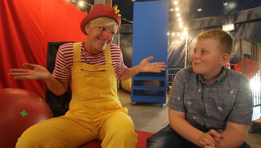 Spaß im Zirkuszelt Links ein Clown, der die Schultern zuckt und einen Jungen, der rechts sitzt, lächelnd anschaut.