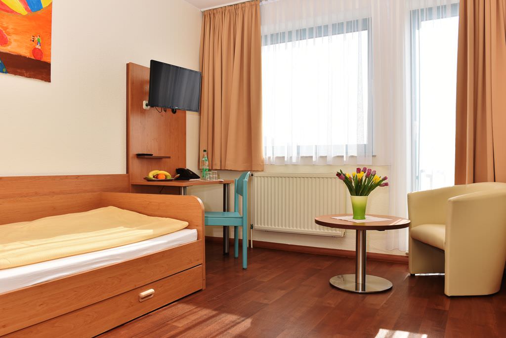 Appartement: links ein Bett. ein Schreibtisch mit Wandfernseher darüber, rechts ein Sessel mit rundem Tisch davor.