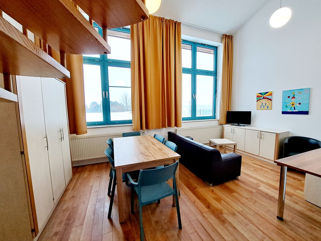 Familienappartement. links in der Mitte ein Holztisch mit sechs Stühlen, recht daneben eine Couch, ganz rechts ein niedriger Schrank mit Fernseher darauf, ganz links ein höherer Schrank und eine Wendeltreppe im Anschnitt.