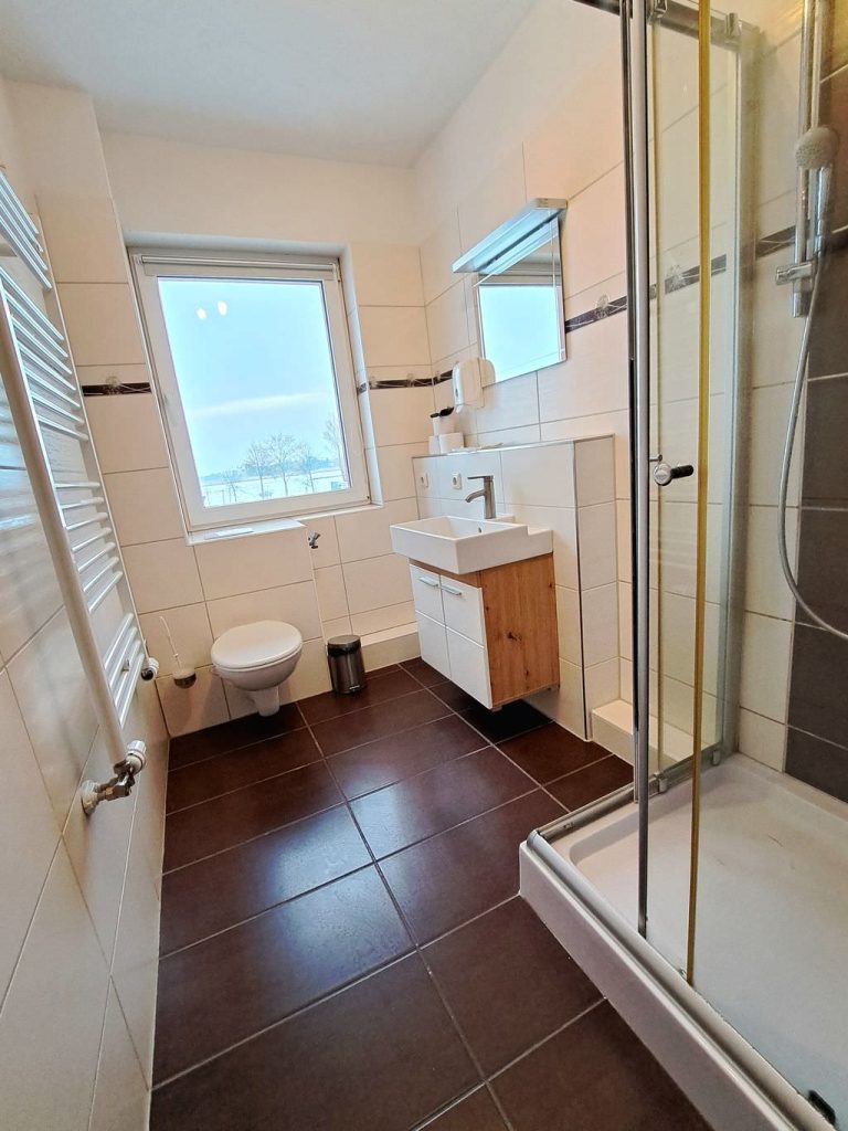 Modernes Badezimmer, hell gefliest, mit Dusche rechts im Vordergrund, dahinter rechts ein Handwaschbecken, in der Mitte vor Kopf ein Fenster, darunter eine Toilette, rechts eine Wandheizung.