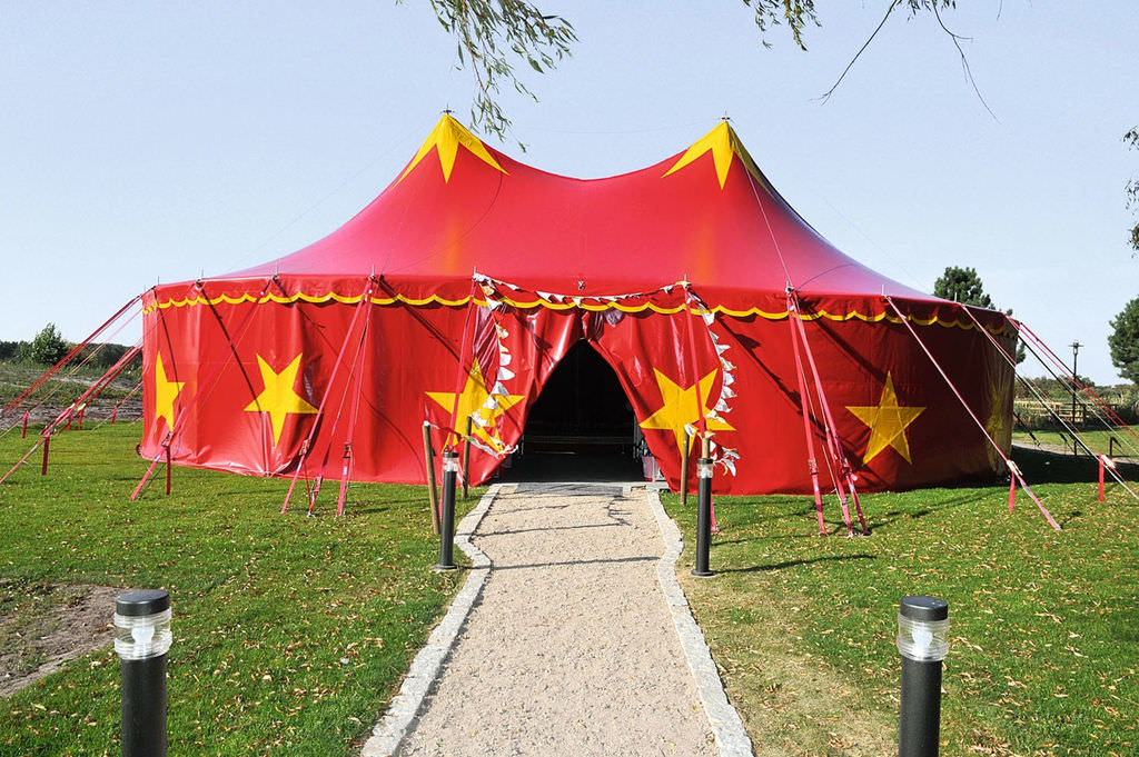 Ein rotes Zirkuszelt mit gelben Sternen und geöffnetem Eingang, auf den ein Fußweg zuführt.