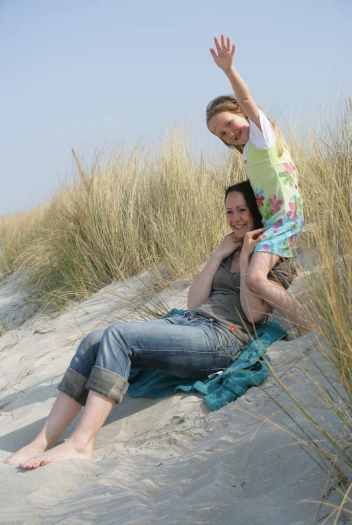 Eine Mutter sitzt im Dünensand vor Gräsern, auf ihrer Schulter ein winkendes Mädchen.
