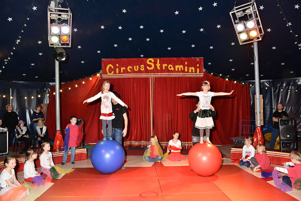 Zwei Mädchen balancieren auf Gymnastikbällen in einer Zirkusmanege umgeben von Kindern, im Hintergrund ein roter Vorhang, über dem Zirkus Stramini steht.