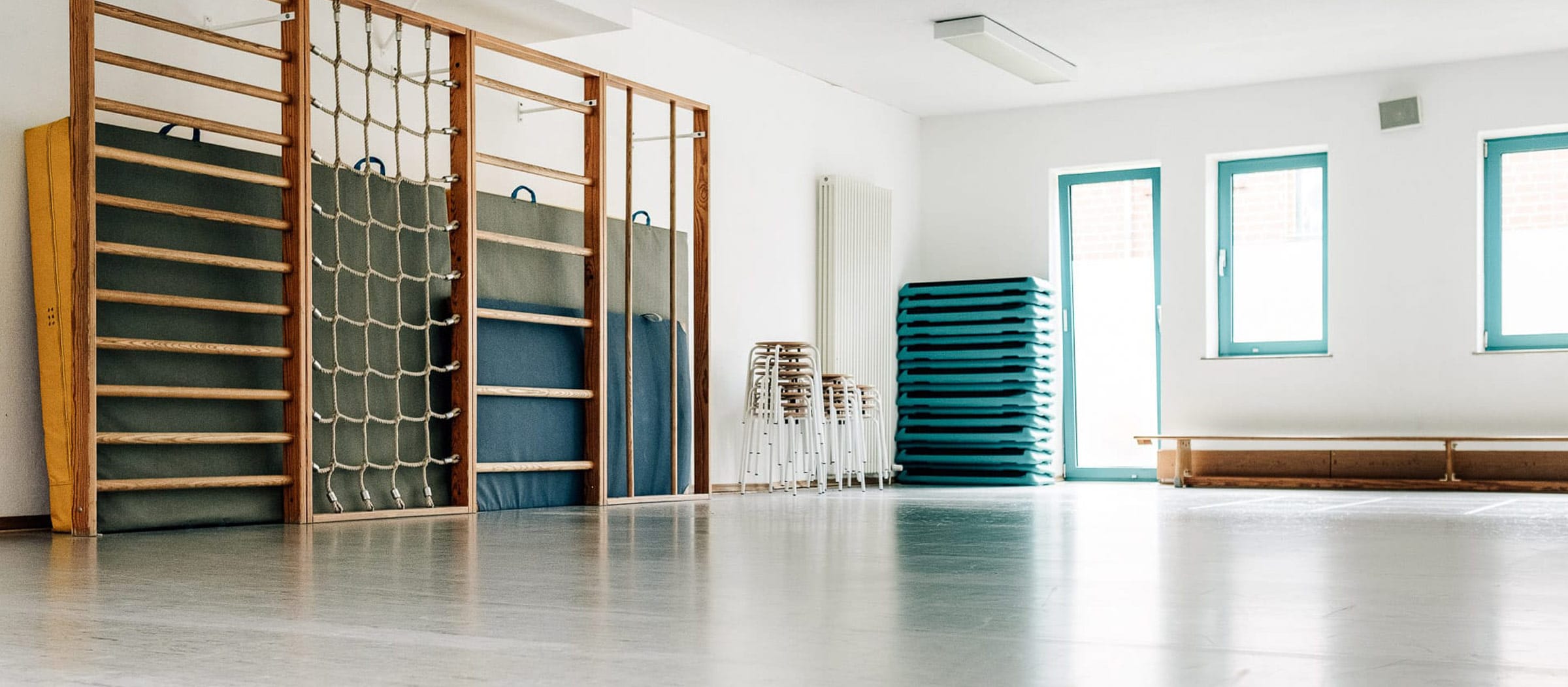 Ein heller Trainingsraum mit Sprossenwand samt Kletterseilnetz links, daneben gestapelte Hocker und Trainingsmatten, rechts vor Kopf eine Holzbank.