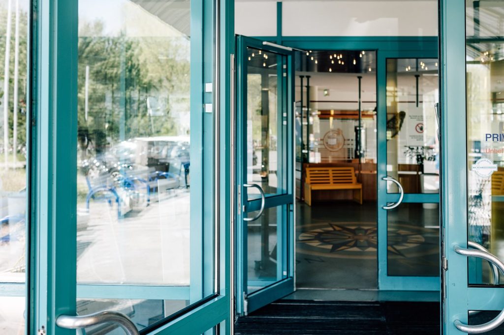 Gläserne, zur Hälfte geöffnete Flügel-Eingangstüre, Blick auf Holzbank im Gebäudeinneren. 