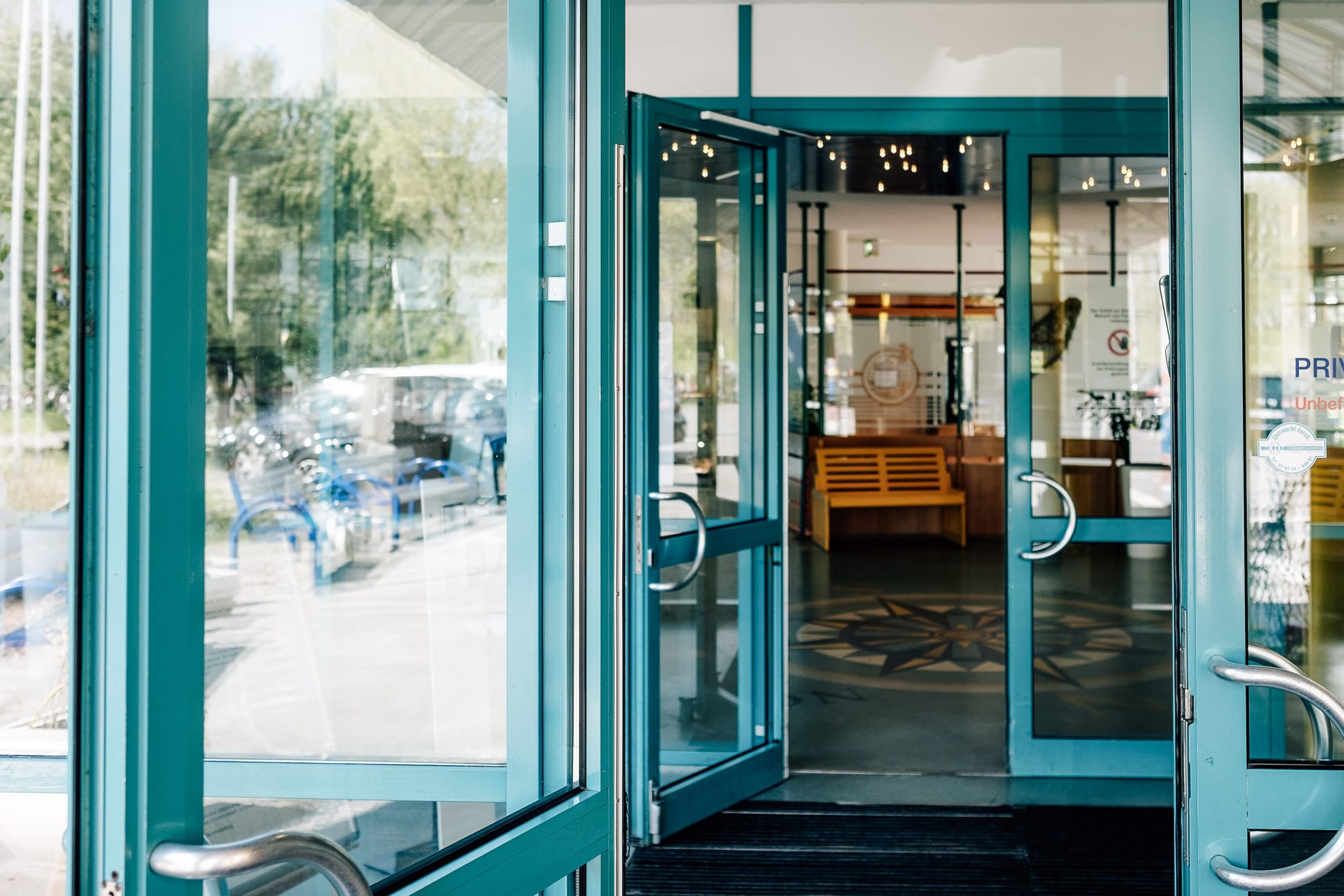 Gläserne, zur Hälfte geöffnete Flügel-Eingangstüre, Blick auf Holzbank im Gebäudeinneren.
