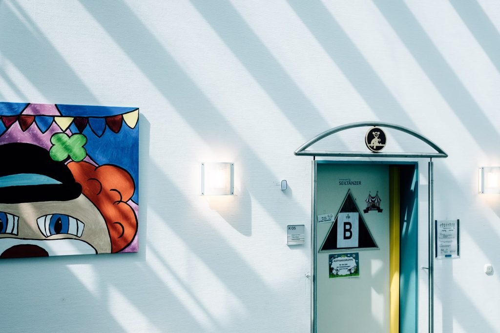 Links das Gemälde eine Clownskopfes an einer weißen Wand, rechts eine Türe, auf der Seiltänzer geschirben steht.