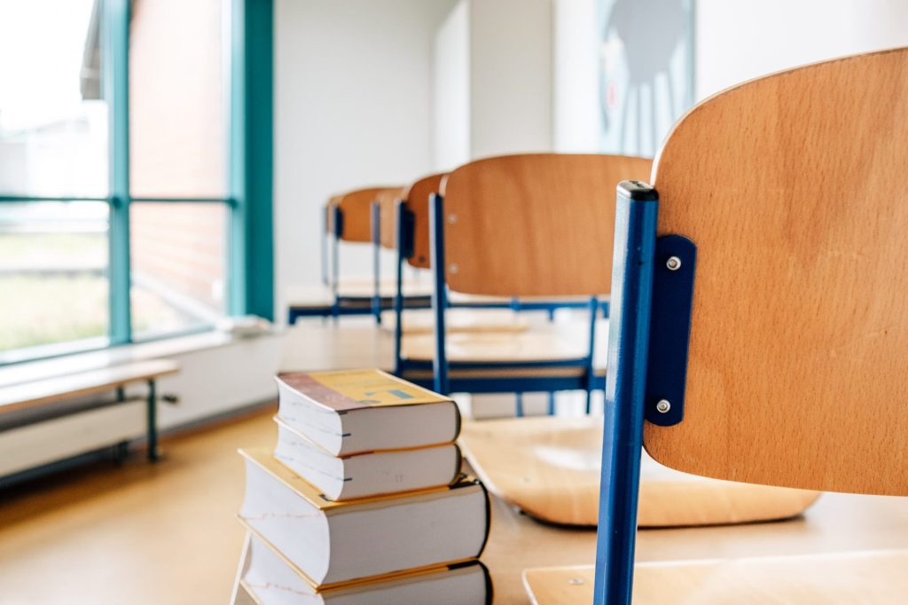 Schultische mit Stühlen daruaf, auf dem Tisch im Vordergrund befinden sich vier gestapelte Schulbücher.