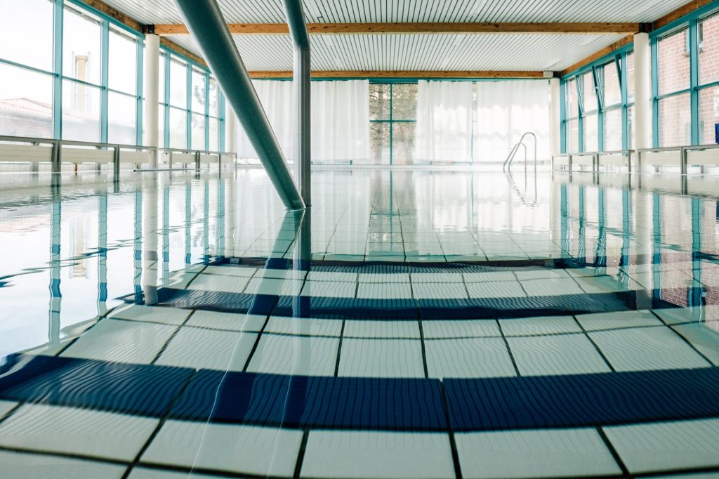 Das helle Schwimm- und Bewegungsbad der Klinik mit weißen und blauen Fliesen auf dem Boden des Beckens, bodentiefe Fenster lassen rundherum Licht herein.