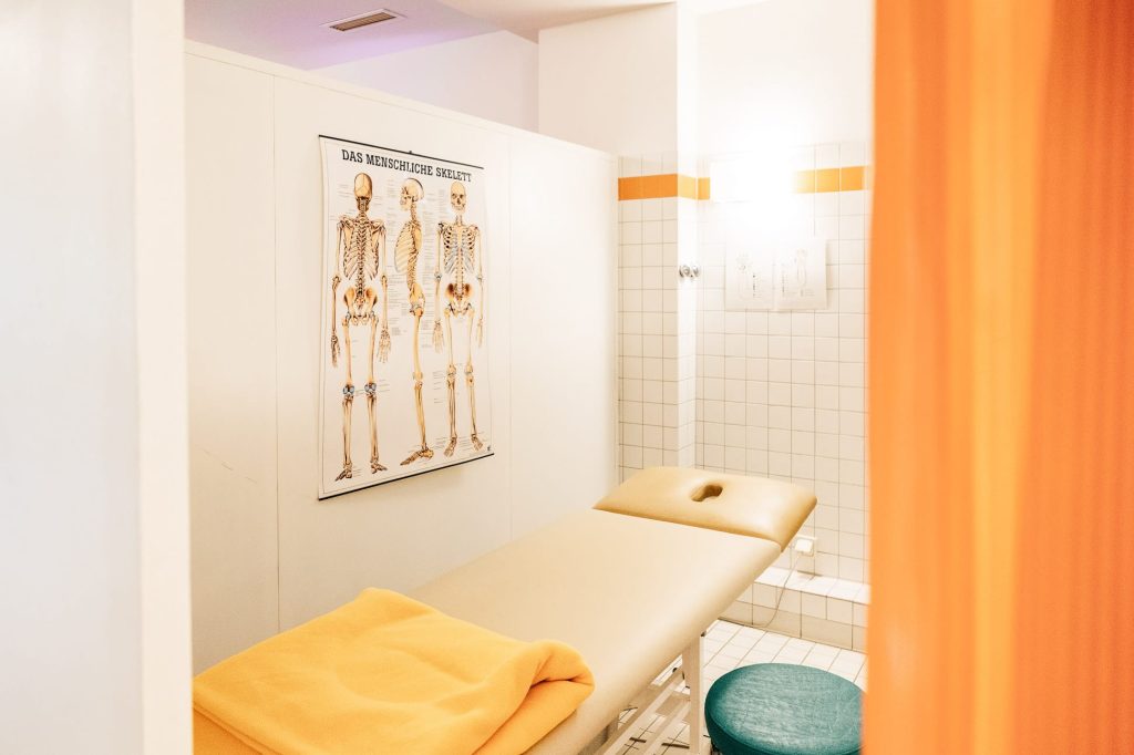 Ein Untersuchungs- und Behandlungszimmer mit weißen Fliesen, links an der Wand das Schaubild eines menschlichen Skeletts, darunter eine Liege.