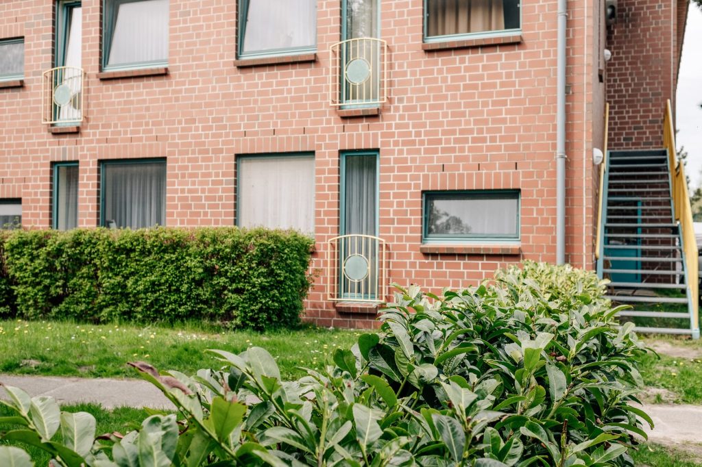 Anschnitt einer Backsteinfassade, rechts davon eine Treppe, davor zwei Bäume.
