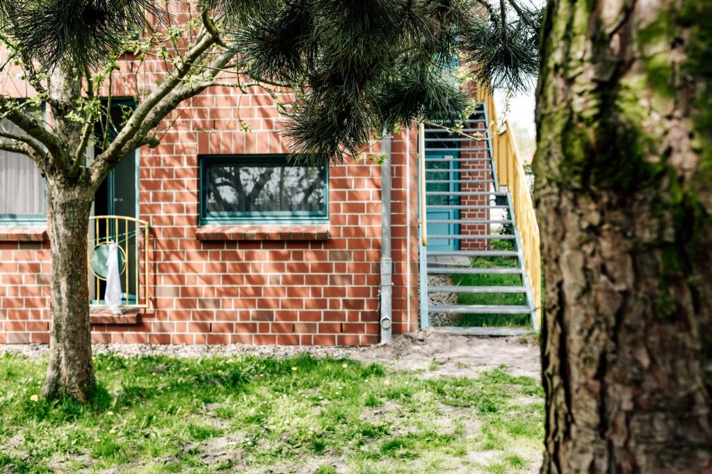 Rechts eine Treppe, die entlang der Fassade eines Backsteingebäudes zu einer Türe führt. Links eine parkänliche Landschaft mit Fußweg.