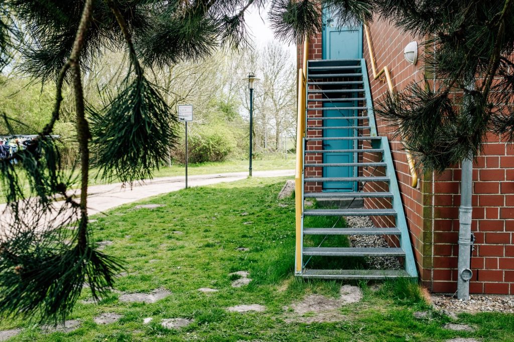Rechts eine Treppe entlang einer Backsteinfassade, die zu einer Türe führt. Links eine Wise mit Fußweg und Bäume.