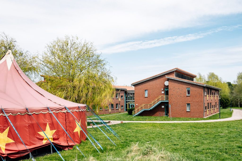 Verschiedene, zweistöckige Backsteingebäude, davor eine Wiese, auf der ein rotes Zirkuszelt mit gelben Sternen links im Anschnitt zu sehen ist.