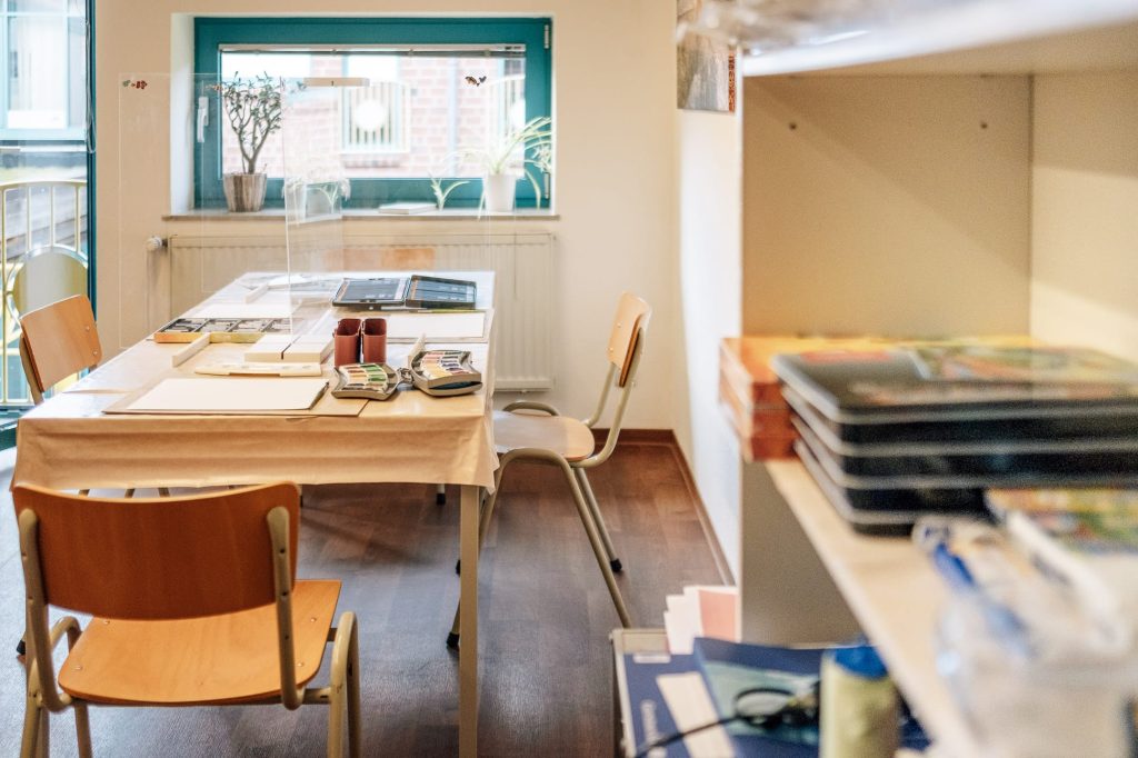 Ein Raum mit einem Tisch, auf dem Malutensilien liegen, rechts ein Regal mit weiteren Utensilien für das kreative Gestalten.